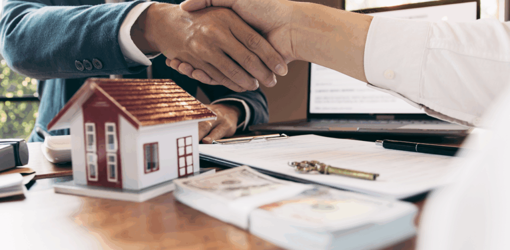 Real estate deal sealed with a handshake beside house model, money, and paperwork