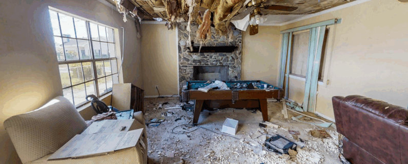 Severely damaged living room with ceiling insulation hanging down, debris on floor, and ruined furniture