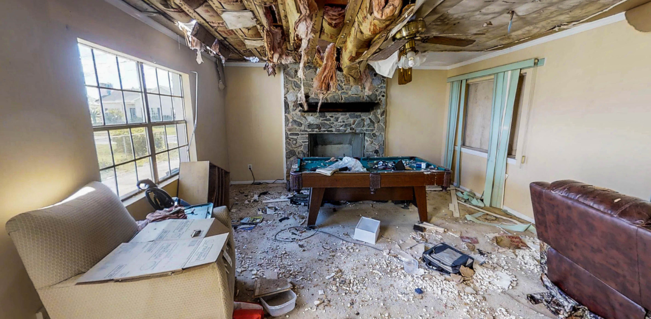 Property Management Severely damaged living room with ceiling insulation hanging down, debris on floor, and ruined furniture
