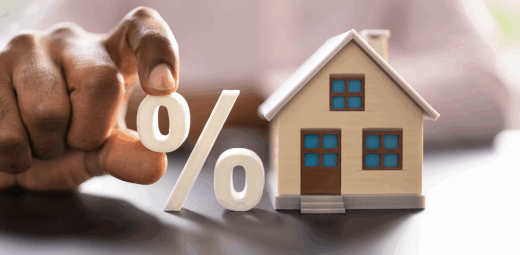 Hand holds a percent sign next to a model house, referencing mortgage interest rates