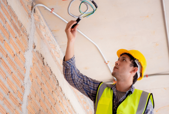 Electrician in hard hat installing wiring through ceiling conduit, highlighting professional electrical services
