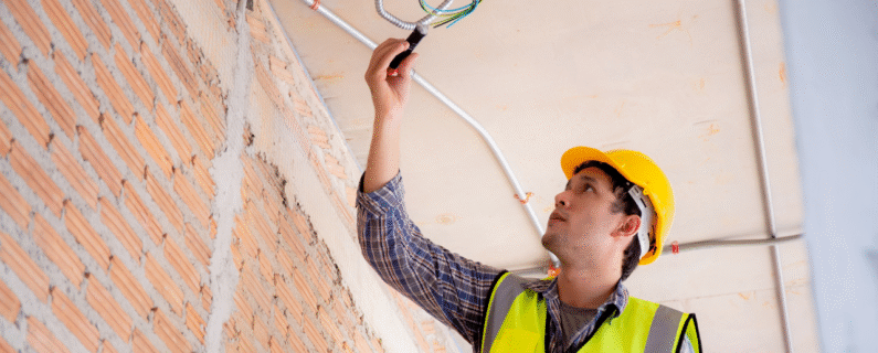 Electrician in hard hat installing wiring through ceiling conduit, highlighting professional electrical services
