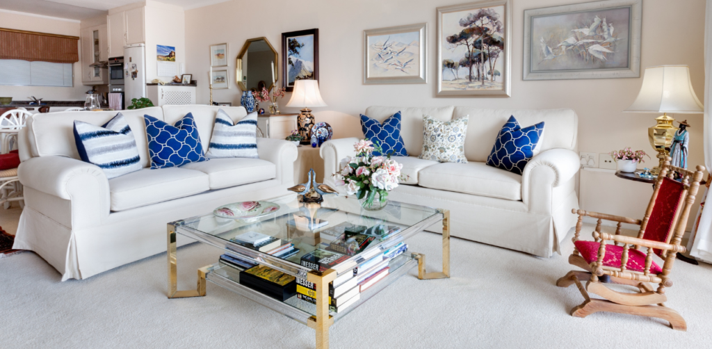 Bright and elegant living room with white sofas, blue and white patterned pillows, a glass coffee table, and framed artwork on the walls