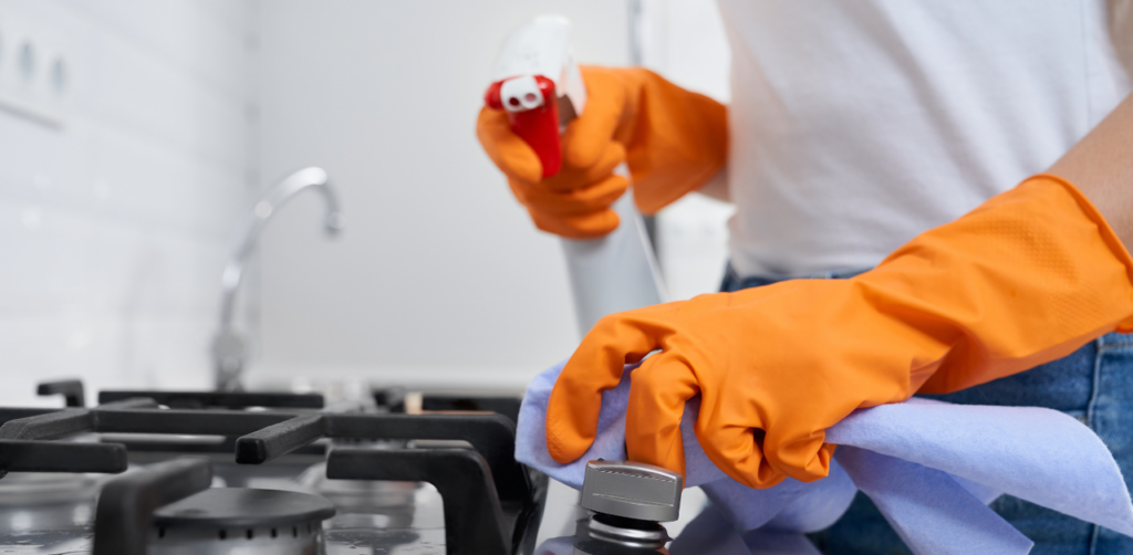 Person wearing orange gloves cleaning a stovetop with a spray bottle and cloth in a kitchen