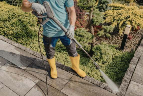Worker pressure washing a stone patio beside landscaped garden beds, wearing gloves and protective rubber boots