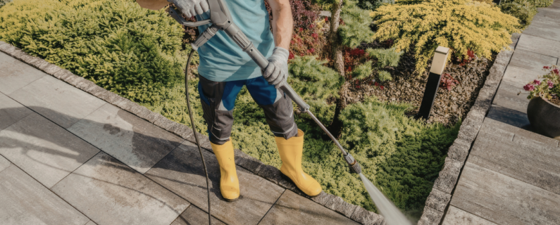 Worker pressure washing a stone patio beside landscaped garden beds, wearing gloves and protective rubber boots