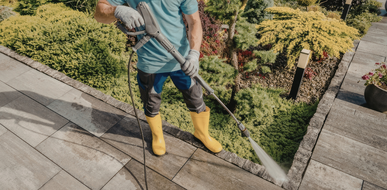 Worker pressure washing a stone patio beside landscaped garden beds, wearing gloves and protective rubber boots