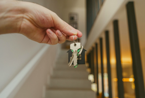 Hand holding a set of house keys with a home-shaped keychain inside a residence, with a staircase visible in the background, symbolizing moving into or purchasing a home