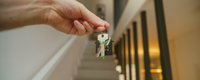 Hand holding a set of house keys with a home-shaped keychain inside a residence, with a staircase visible in the background, symbolizing moving into or purchasing a home