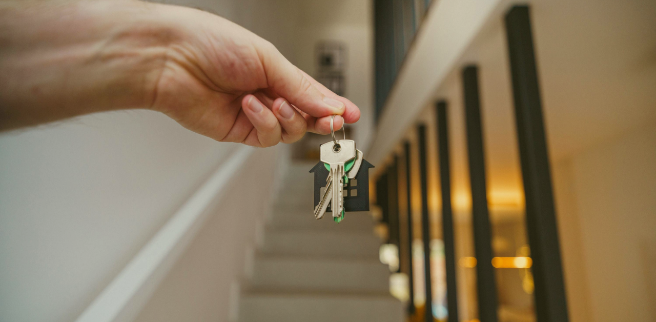 Hand holding a set of house keys with a home-shaped keychain inside a residence, with a staircase visible in the background, symbolizing moving into or purchasing a home