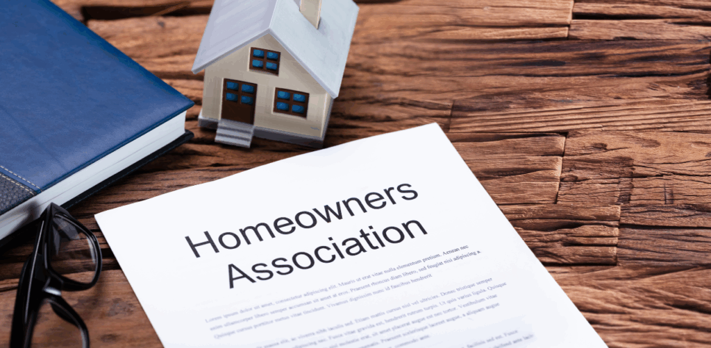 Homeowners association document sits on a rustic wooden table alongside a small house model, glasses, and a book, representing community rules or residential legal agreements