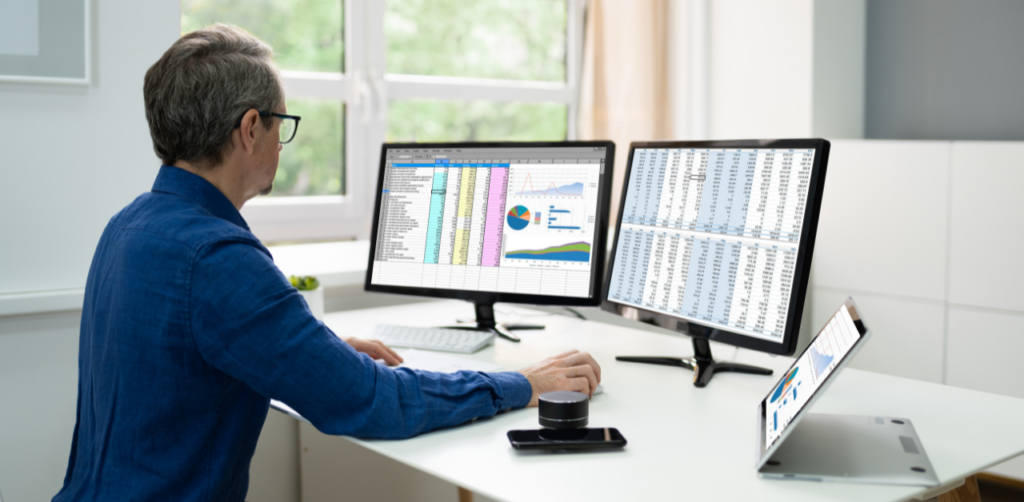 Man in a blue shirt working on spreadsheets and graphs displayed across multiple monitors in a modern office setting