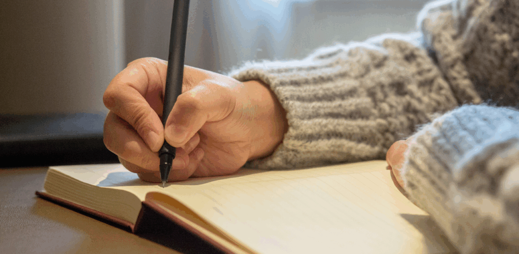 Person in a cozy sweater writes in a blank notebook with a black pen, possibly taking notes or drafting a legal statement