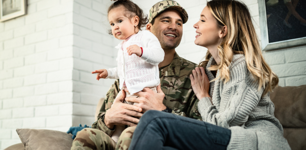 Soldier in uniform sitting on a couch with his family, holding a young child while smiling at his partner