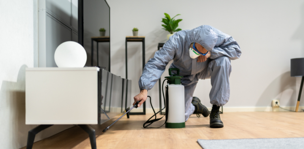 A pest control worker in protective gear spraying beneath furniture inside a modern living space