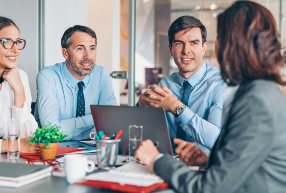 Four professionals in business attire sit at a table during a meeting, with laptops, notebooks, and water glasses visible