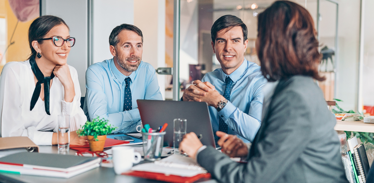 Four professionals in business attire sit at a table during a meeting, with laptops, notebooks, and water glasses visible