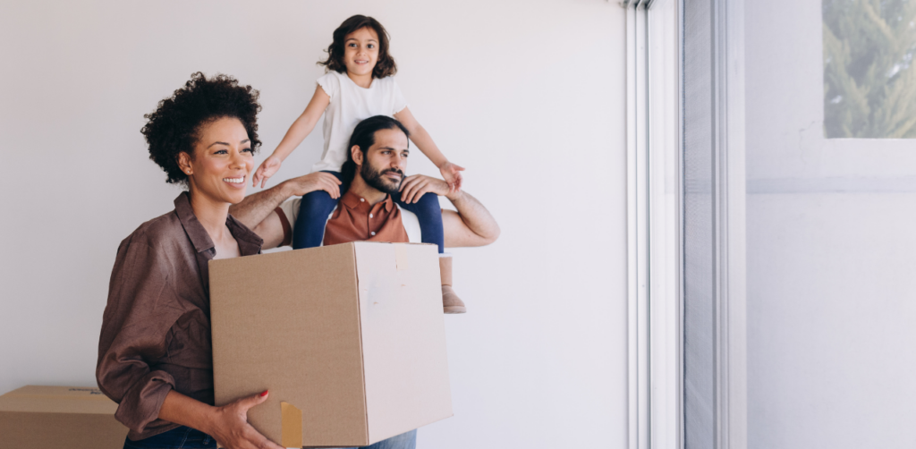 A smiling family moving into a new home, with the woman holding a box and a man carrying a child on his shoulders