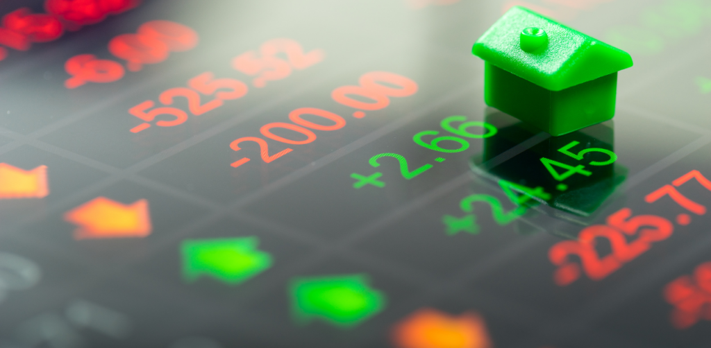 A green toy house sits on a digital stock display showing fluctuating numbers in red and green, symbolizing real estate market changes