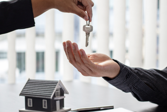 A hand reaches out to pass a key to another open hand over a table with a small house model in the background