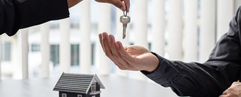 A hand reaches out to pass a key to another open hand over a table with a small house model in the background