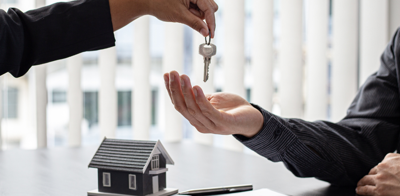 A hand reaches out to pass a key to another open hand over a table with a small house model in the background