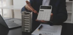 A person in business attire holds a large calculator displaying numbers and points at a model building, with contract papers and a laptop on the table