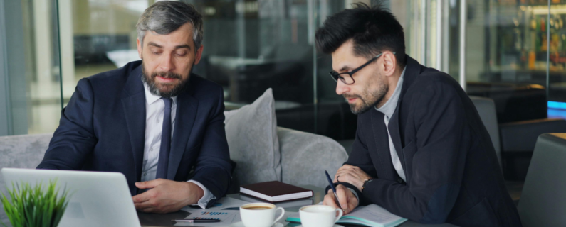 Two business professionals in suits reviewing data on a laptop over coffee, collaborating on financial planning