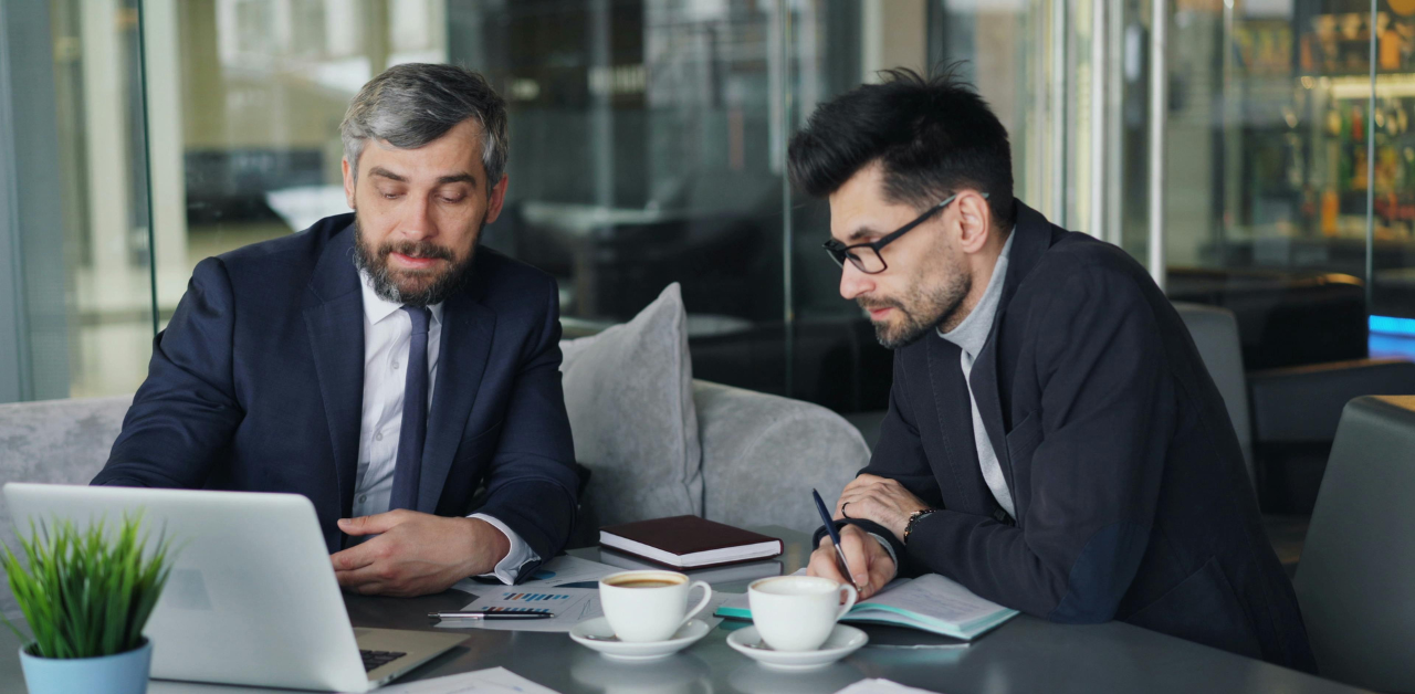 Two business professionals in suits reviewing data on a laptop over coffee, collaborating on financial planning