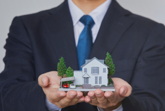 Person in a business suit holding a miniature model house with both hands, symbolizing home ownership, real estate, or insurance