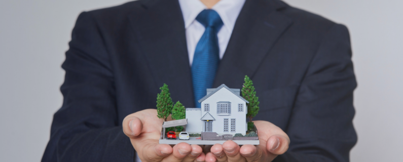 Person in a business suit holding a miniature model house with both hands, symbolizing home ownership, real estate, or insurance