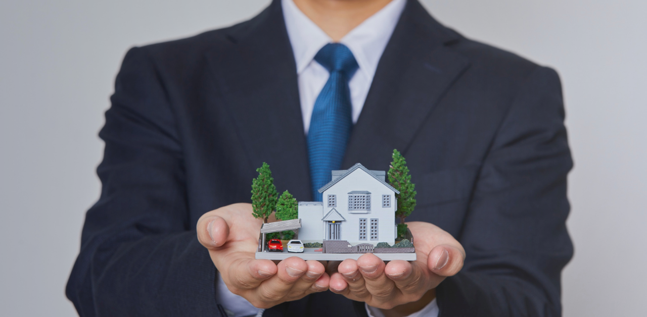 Person in a business suit holding a miniature model house with both hands, symbolizing home ownership, real estate, or insurance