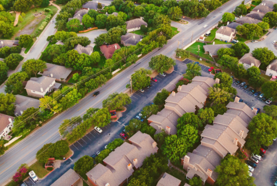 Aerial view of a tree-lined suburban neighborhood with houses, apartment buildings, parking lots, and a main road