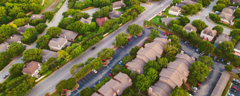 Aerial view of a tree-lined suburban neighborhood with houses, apartment buildings, parking lots, and a main road