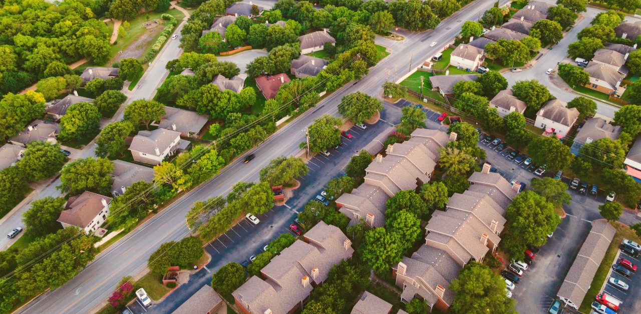 Aerial view of a tree-lined suburban neighborhood with houses, apartment buildings, parking lots, and a main road